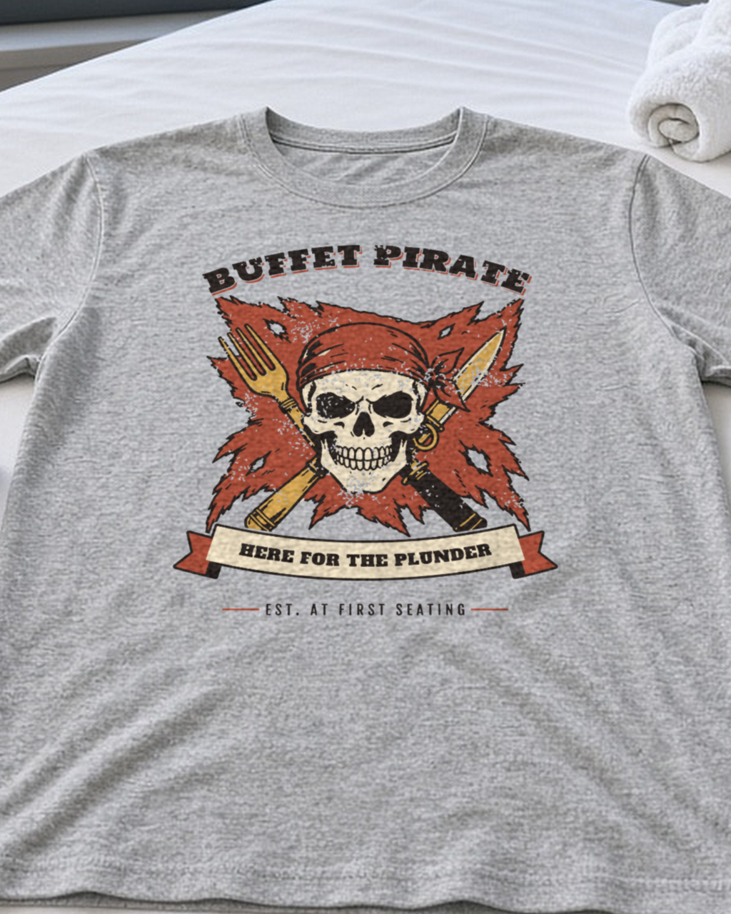 Mockup: ash Buffet Pirate tee laid flat on a cabin bed with a towel animal and pirate hat